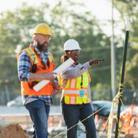 Two multiracial construction workers or engineers walking side by side through a construction site, conversing. One of them is a mature African-American woman in her 40s. Piles of dirt are in the foreground and a road is in the background.
